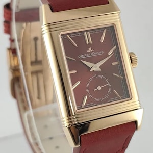 May include: A gold-toned rectangular wristwatch with a burgundy face and a small seconds dial. The watch features gold hands and hour markers, and a burgundy leather strap. The watch face has the "Jaeger-LeCoultre" logo.