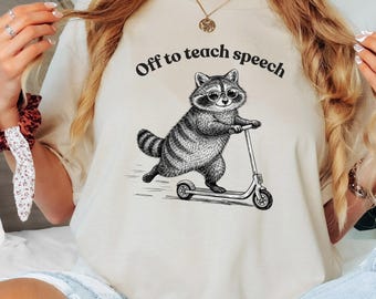 May include: Beige t-shirt with a graphic of a raccoon wearing glasses and riding a scooter. The text "Off to teach speech" is arched above the raccoon. The design is in black and white.