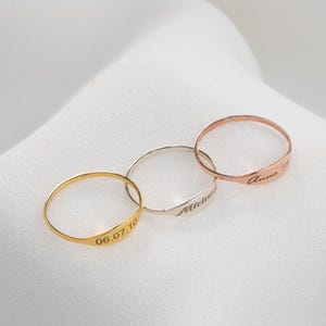 Könnte beinhalten: Drei zarte Ringe in Gold, Silber und Roségold, jeweils mit einer einzigartigen Inschrift graviert. Der goldene Ring zeigt das Datum "06.07.10", der silberne Ring hat den Namen "Michael" und der roségoldene Ring trägt die Aufschrift "Anna". Die Ringe werden auf einer weißen, strukturierten Oberfläche präsentiert.