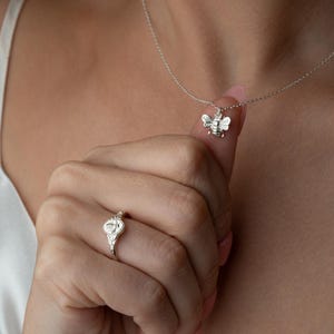 May include: A silver bee pendant necklace and a silver ring with a floral design. The bee pendant is small and detailed, and the ring has a delicate, ornate appearance. The jewellery is displayed against a soft background.