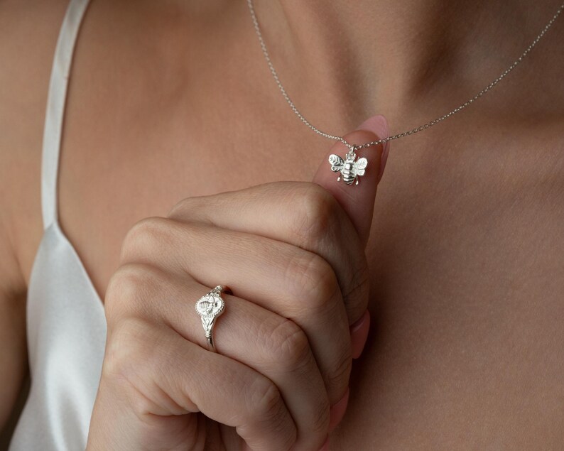 May include: A silver bee pendant necklace and a silver ring with a floral design. The bee pendant is small and detailed, and the ring has a delicate, ornate appearance. The jewellery is displayed against a soft background.