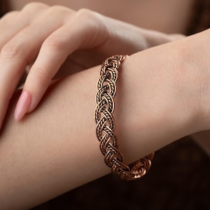 May include: A copper-colored braided bracelet is worn on a wrist. The bracelet has a detailed woven design. The warm tones of the bracelet contrast with the skin tone. The bracelet is a piece of jewelry.