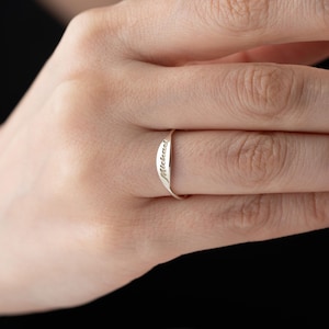 May include: A slender, silver ring with the name "Michael" engraved on the band. The ring is worn on a finger, with the hand and a dark background visible.