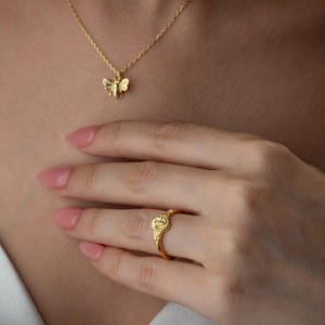 May include: A gold-coloured bee pendant necklace and a gold ring with a decorative design. The ring is on a person's finger. The necklace and ring are likely made of gold or a gold-coloured metal.