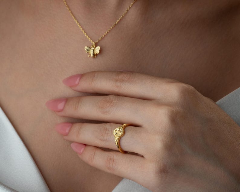 May include: A gold-coloured bee pendant necklace and a gold ring with a decorative design. The ring is on a person's finger. The necklace and ring are likely made of gold or a gold-coloured metal.