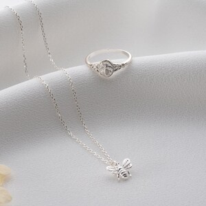 May include: A silver bee pendant necklace and a matching bee ring are presented on a white surface. The ring has a bee design at its centre. The necklace features a fine chain and a detailed bee pendant.
