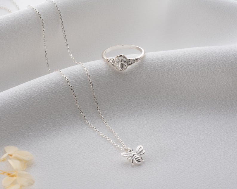 May include: A silver bee pendant necklace and a matching bee ring are presented on a white surface. The ring has a bee design at its centre. The necklace features a fine chain and a detailed bee pendant.