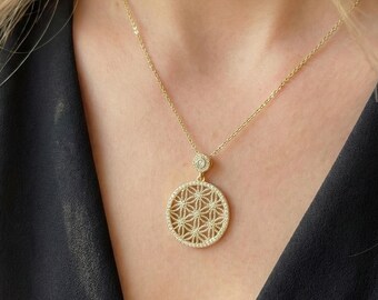 May include: A gold-colored necklace with a circular pendant featuring a geometric floral design. The pendant is encrusted with small, clear gemstones. The necklace is worn against a black top.