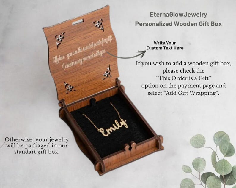 May include: A wooden gift box with a personalized necklace inside. The box is open, revealing a gold-coloured necklace spelling "Emily" on a black velvet interior. The box has decorative cutouts and engraved text.