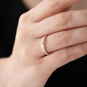 Op de afbeelding: Een gouden ring met een rechthoekige, lichtroze edelsteen. De ring heeft een minimalistisch ontwerp en zit om een vinger. De achtergrond is zwart.