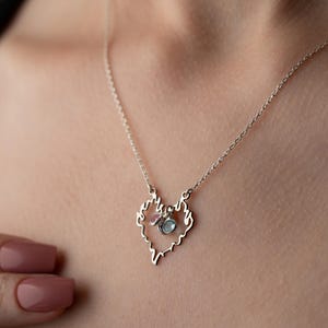 May include: A delicate silver necklace featuring a heart-shaped pendant with a textured outline. The pendant is adorned with small pink and blue gemstones. The necklace is displayed on a person's neck.