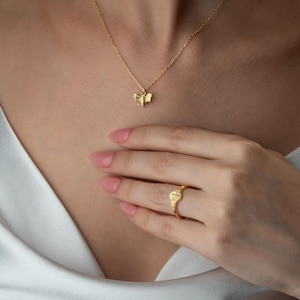 May include: A gold necklace with a bee pendant and a matching ring. The ring has a heart-shaped design. The jewellery is worn against a white satin top.
