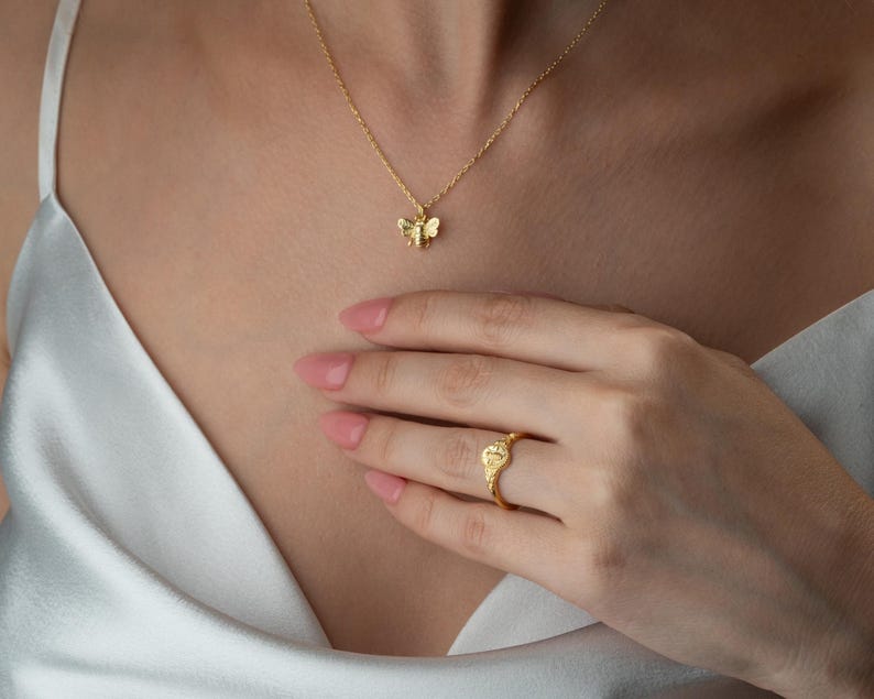 May include: A gold necklace with a bee pendant and a matching ring. The ring has a heart-shaped design. The jewellery is worn against a white satin top.