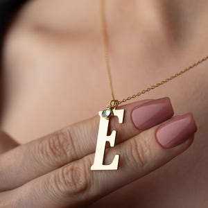 May include: A gold-colored initial pendant necklace with the letter "E". The pendant has a small, light-green gemstone. The necklace is on a delicate chain. The background is blurred, focusing on the necklace.