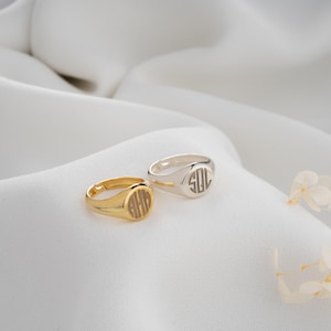 Könnte beinhalten: Zwei Siegelringe, einer goldfarben und einer silberfarben, liegen auf einer weißen, strukturierten Oberfläche. Der goldfarbene Ring zeigt die Initialen "AHG" in einem kreisförmigen Design. Der silberfarbene Ring zeigt die Initialen "SOL". Beide Ringe haben eine klassische, runde Form.