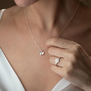 May include: A silver bee pendant necklace and a silver ring with a heart-shaped design. The necklace is delicate, and the ring features small, sparkling stones. The jewelry is worn against a light-coloured top.