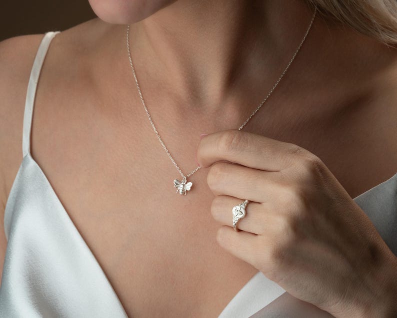 May include: A silver bee pendant necklace and a silver ring with a heart-shaped design. The necklace is delicate, and the ring features small, sparkling stones. The jewelry is worn against a light-coloured top.