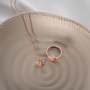 May include: A rose gold bee pendant necklace and ring set. The necklace has a fine chain and a detailed bee pendant. The ring has a similar design with a small gemstone. Both items are displayed on a decorative plate.