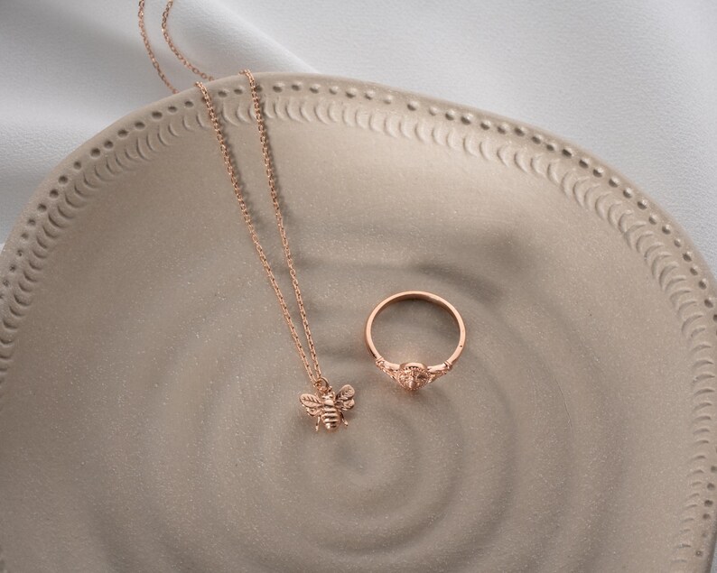 May include: A rose gold bee pendant necklace and ring set. The necklace has a fine chain and a detailed bee pendant. The ring has a similar design with a small gemstone. Both items are displayed on a decorative plate.