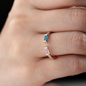 Peut inclure: Une bague dorée de conception ouverte, ornée d'une pierre précieuse rectangulaire bleue et rose. La bague est portée au doigt, mettant en valeur les couleurs des pierres et le style minimaliste de la bague.