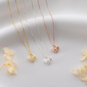 May include: Four delicate bee pendant necklaces in gold, silver, and rose gold. The bee pendants are detailed, and the chains are thin. The necklaces are displayed on a white, textured surface with dried flowers.