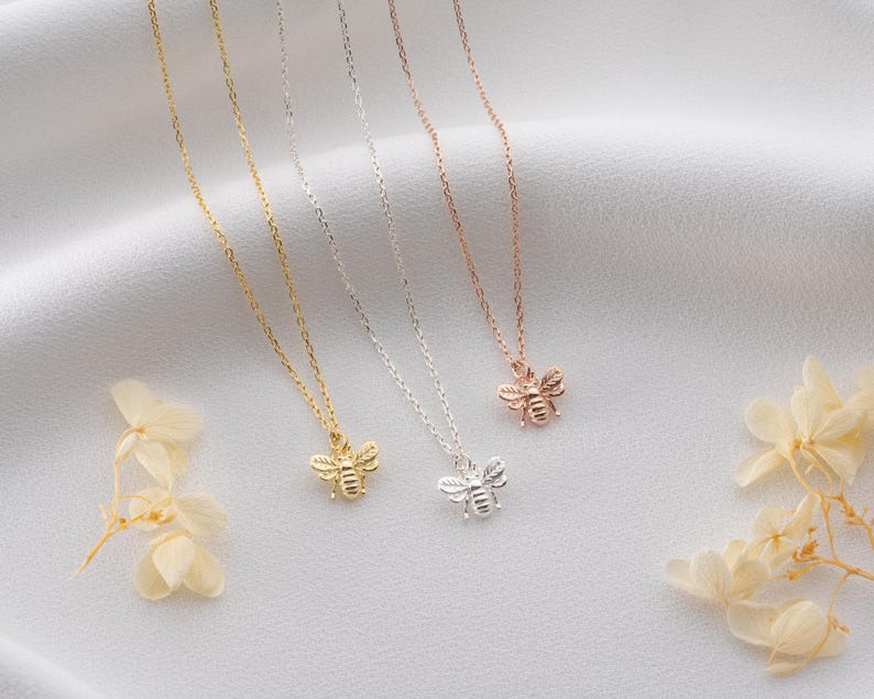 May include: Four delicate bee pendant necklaces in gold, silver, and rose gold. The bee pendants are detailed, and the chains are thin. The necklaces are displayed on a white, textured surface with dried flowers.
