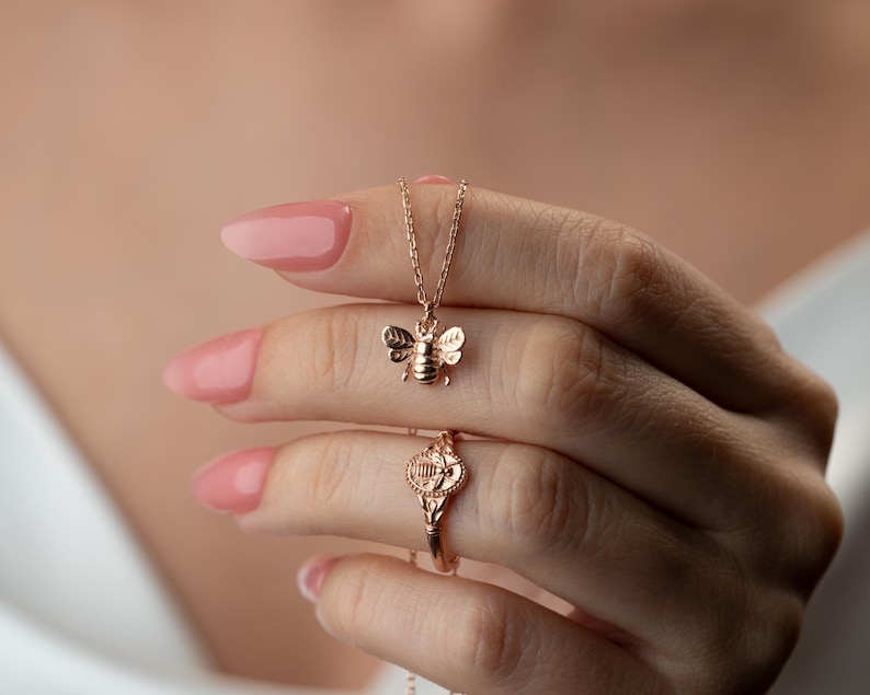 May include: A gold-coloured bee pendant necklace and a matching ring. The bee pendant is detailed with wings and body segments. The ring features a similar design with an ornate, circular emblem. The jewellery is displayed against a soft, blurred background.