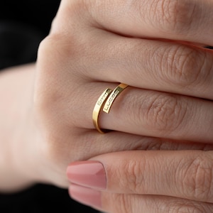 May include: A gold-colored ring with the names "Emma" and "Lucas" engraved on it. The ring has a unique, open design where the band wraps around the finger. The ring is shown on a hand with a pink nail polish.