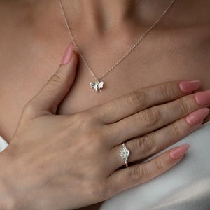 May include: A silver bee pendant necklace and a silver ring with a floral design. The ring is on the ring finger of a hand with pink nail varnish. The necklace is on a person's neck, and the hand is resting on the person's chest.