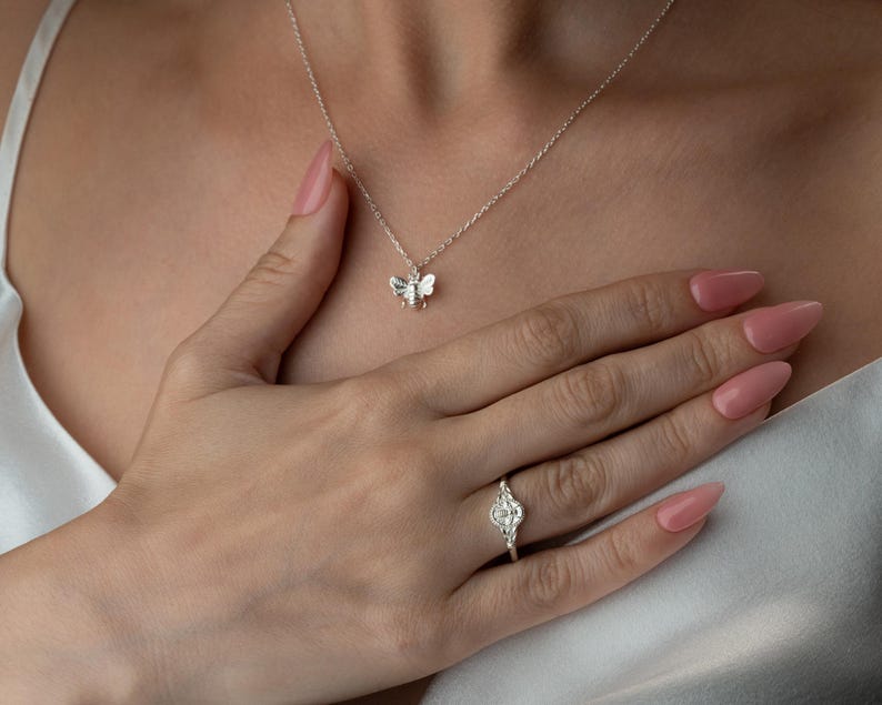 May include: A silver bee pendant necklace and a silver ring with a floral design. The ring is on the ring finger of a hand with pink nail varnish. The necklace is on a person's neck, and the hand is resting on the person's chest.