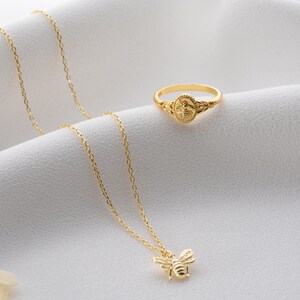 May include: A gold ring and necklace set. The ring has a detailed design, and the necklace features a bee pendant. Both items are displayed on a white, textured surface, highlighting the jewellery's intricate details and shine.