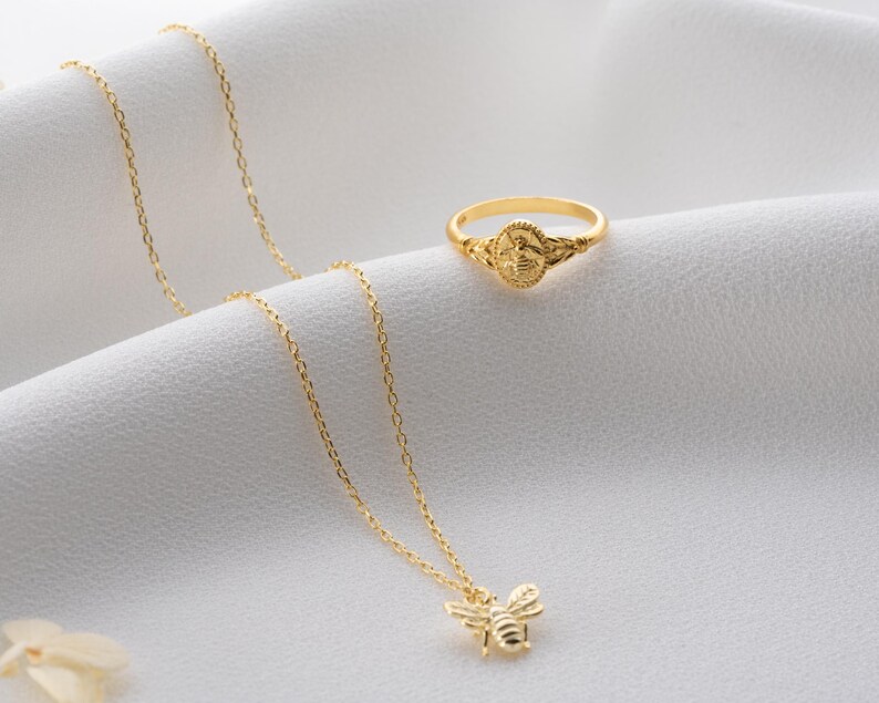 May include: A gold ring and necklace set. The ring has a detailed design, and the necklace features a bee pendant. Both items are displayed on a white, textured surface, highlighting the jewellery's intricate details and shine.