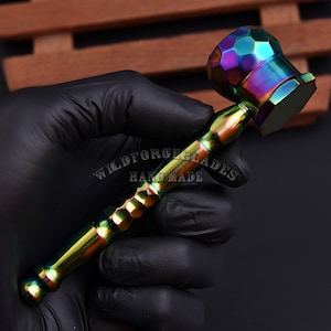 Puede incluir: Un martillo iridiscente multicolor con cabeza y mango facetados. El mango tiene un patrón hexagonal texturizado. El texto "WILDFORGEBLADES HAND MADE" es visible en el mango.