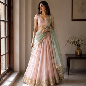 May include: A pale pink lehenga set featuring a short-sleeved blouse with a sweetheart neckline and a floor-length pleated skirt. The ensemble includes a sheer sage green dupatta draped over the shoulder, accented with gold embroidered borders.