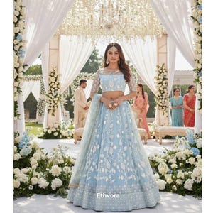 May include: A light blue lehenga with intricate white floral embroidery. The outfit features a cropped top with sheer sleeves and a matching skirt. The scene is set at an outdoor wedding with floral decorations and a decorated canopy.