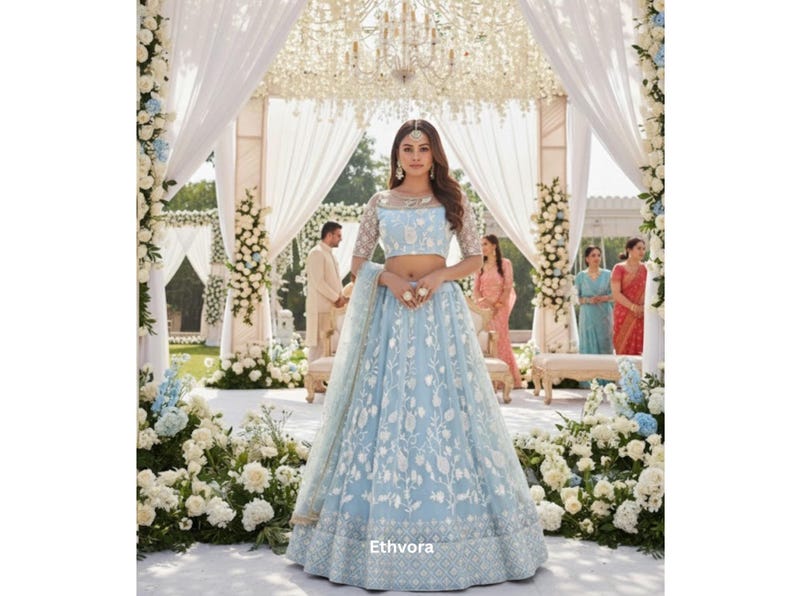 May include: A light blue lehenga with intricate white floral embroidery. The outfit features a cropped top with sheer sleeves and a matching skirt. The scene is set at an outdoor wedding with floral decorations and a decorated canopy.