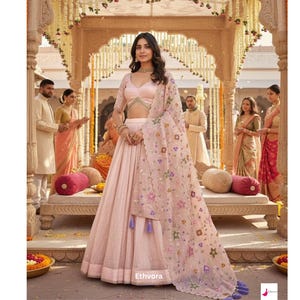 May include: A woman in a pale pink lehenga, blouse, and a sheer dupatta with floral embroidery. The outfit has a flowing skirt and is accessorized with jewellery. The background features an ornate structure and several people.
