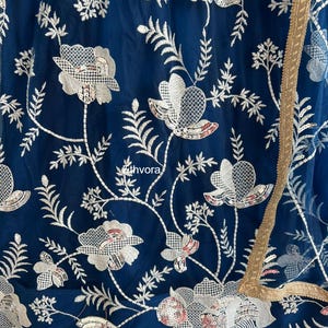 May include: Close-up of a navy blue fabric with intricate white floral embroidery and sequin details. The design includes stylized flowers and leaves, with a gold trim along the edge. The fabric has a sheer quality.