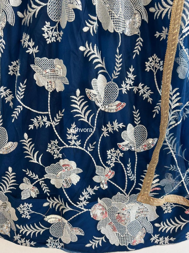 May include: Close-up of a navy blue fabric with intricate white floral embroidery and sequin details. The design includes stylized flowers and leaves, with a gold trim along the edge. The fabric has a sheer quality.