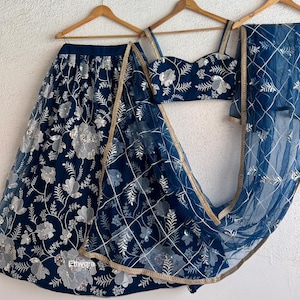 May include: A navy blue embroidered lehenga set with silver floral patterns. The set includes a skirt, a crop top, and a sheer dupatta. The dupatta has a gold trim. The outfit is displayed on wooden hangers.