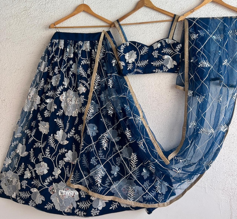 May include: A navy blue embroidered lehenga set with silver floral patterns. The set includes a skirt, a crop top, and a sheer dupatta. The dupatta has a gold trim. The outfit is displayed on wooden hangers.