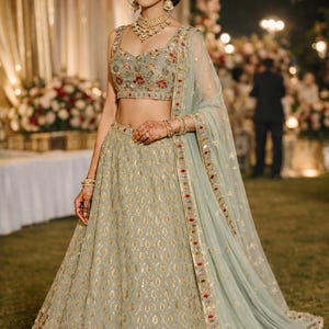 May include: A sage green lehenga set featuring a sleeveless blouse and a floor-length skirt, both adorned with intricate gold embroidery, floral patterns in red and blue, and sequin detailing. A matching sheer dupatta with a decorative border.