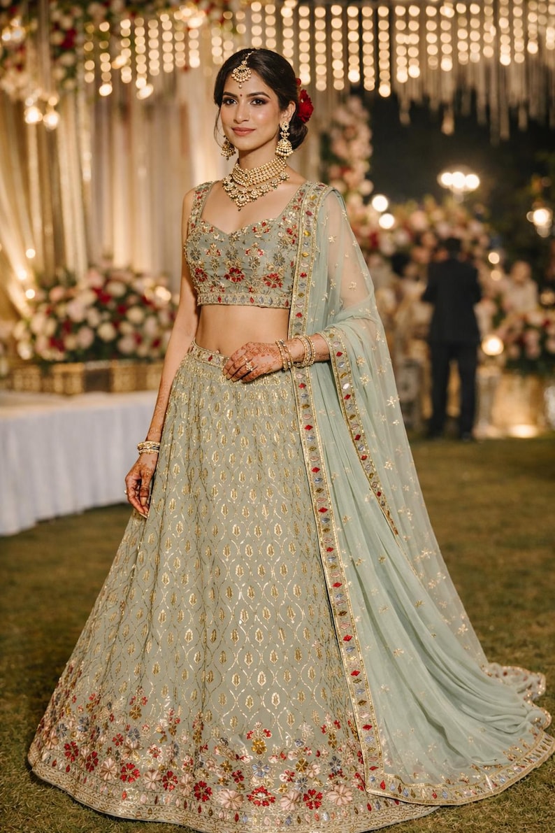 May include: A sage green lehenga set featuring a sleeveless blouse and a floor-length skirt, both adorned with intricate gold embroidery, floral patterns in red and blue, and sequin detailing. A matching sheer dupatta with a decorative border.