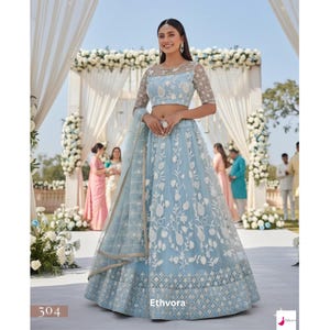May include: A light blue lehenga with white floral embroidery. The outfit includes a matching sheer dupatta and a cropped top with sheer sleeves. The lehenga features a detailed geometric pattern at the hem. The brand name "Ethvora" is visible.