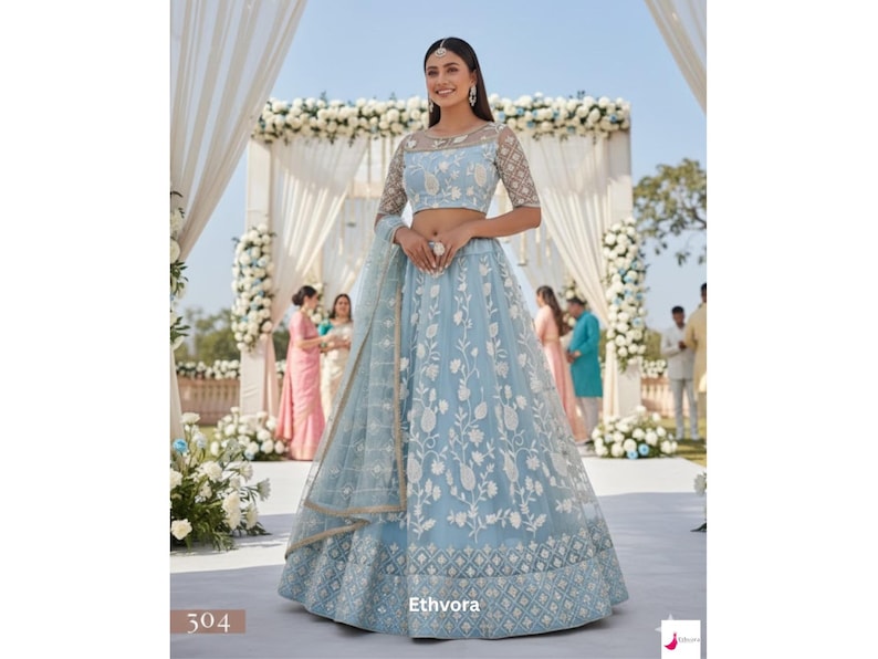 May include: A light blue lehenga with white floral embroidery. The outfit includes a matching sheer dupatta and a cropped top with sheer sleeves. The lehenga features a detailed geometric pattern at the hem. The brand name "Ethvora" is visible.