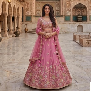 Peut inclure: Une lehenga choli rose, tenue indienne traditionnelle, avec des broderies dorées complexes. L'ensemble comprend un dupatta assorti et un haut sans manches. Le modèle porte un collier et un bracelet. L'arrière-plan est une cour avec des détails architecturaux.