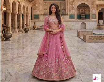 Peut inclure: Une lehenga choli rose, tenue indienne traditionnelle, avec des broderies dorées complexes. L'ensemble comprend un dupatta assorti et un haut sans manches. Le modèle porte un collier et un bracelet. L'arrière-plan est une cour avec des détails architecturaux.