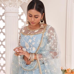May include: A light blue embroidered formal dress with a sheer overlay and gold trim. The dress features floral embroidery and is accessorized with gold jewelry, including earrings, bracelets, and rings. The image also shows a matching sheer scarf.