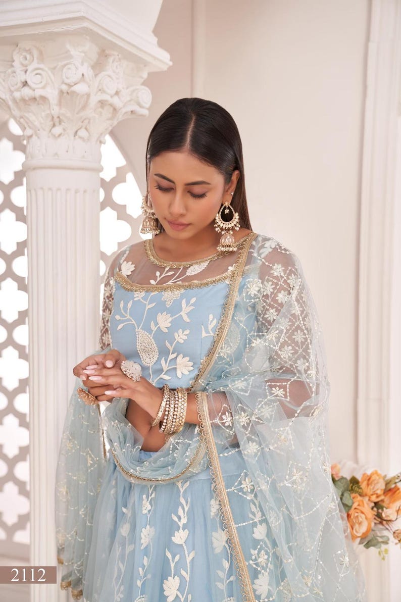 May include: A light blue embroidered formal dress with a sheer overlay and gold trim. The dress features floral embroidery and is accessorized with gold jewelry, including earrings, bracelets, and rings. The image also shows a matching sheer scarf.