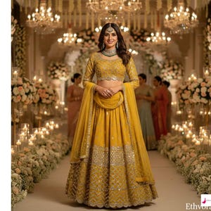 Op de afbeelding: Een vrouw in een mosterdgele lehenga choli, een traditionele Indiase outfit, staat in een versierde hal. De outfit heeft ingewikkelde borduursels en een bijpassende dupatta. De achtergrond omvat bloemstukken, kaarsen en kroonluchters.